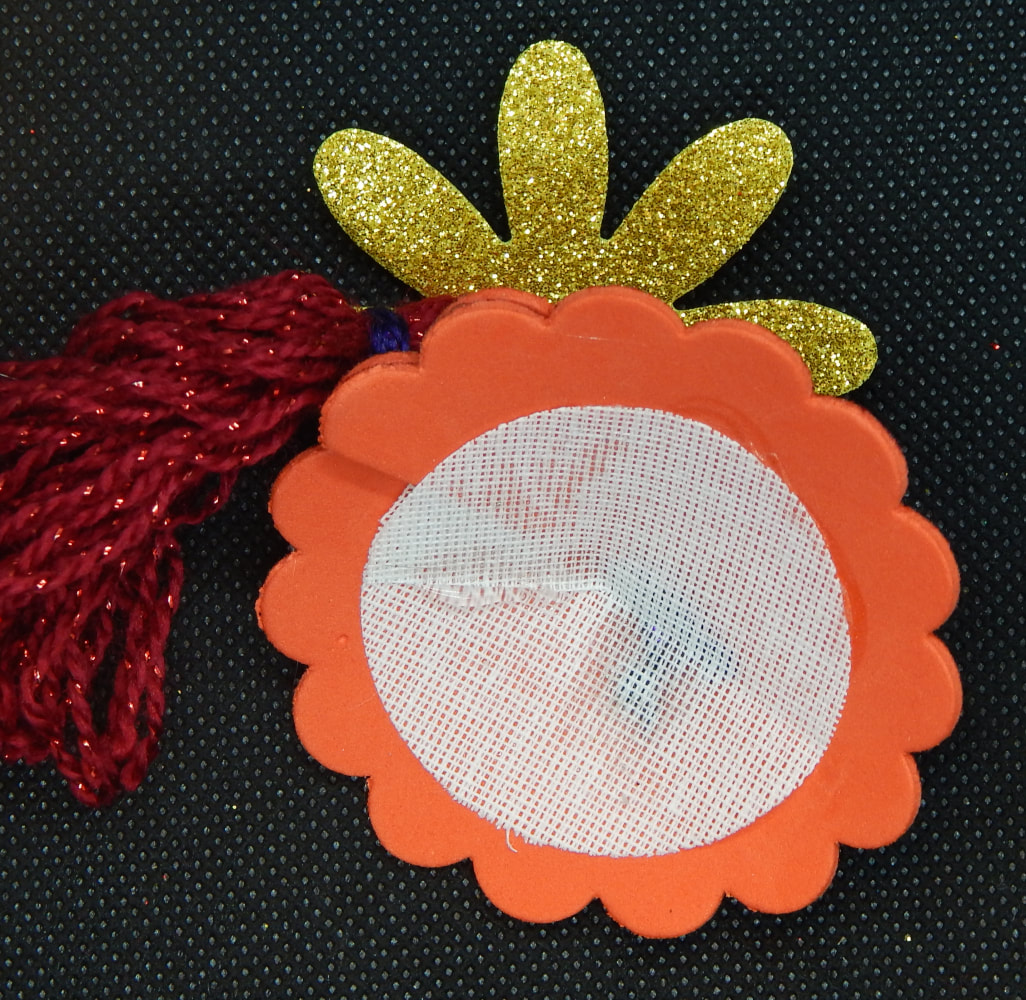 A picture of the back of the in-progress pastie. The white buckram fabric circle is completely glued down on the red circle of craft foam with the scallops. A red tassel with a purple tie can be seen off to the left, and a gold glitter craft foam flower edge is peeking out from the top. 