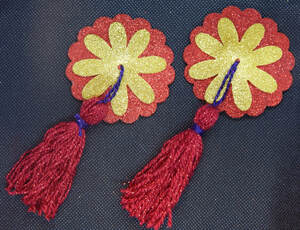 A pair of pasties (nipple covers) with a red circle base, yellow flower, and red and purple tassel lie diagonally on a black background 