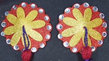 A close up of the pasties (nipple covers) They have a red glitter background and a yellow glitter flower shape on top. There are silver/clear gems glued on around the scalopped edge. Just the purple cord and very top of a red tassel can be seen coming from the center. 