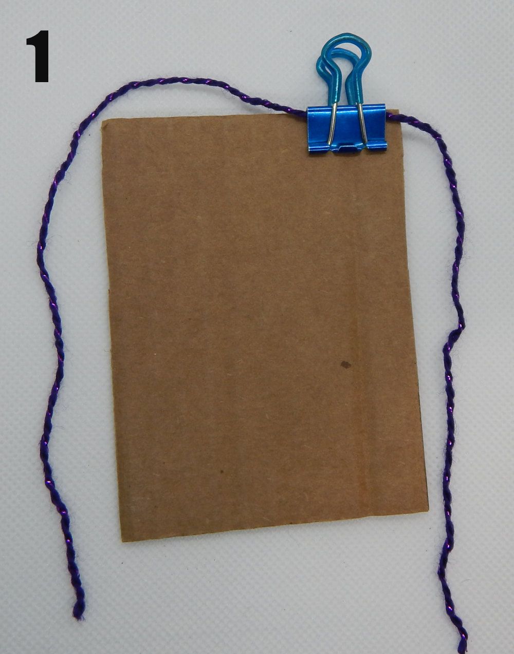 A rectangle of brown cardboard is in the middle. There is a blue binder clip on the upper right corner holding a piece of purple thread onto the cardboard. There is a 1 in the upper left hand corner of the picture.