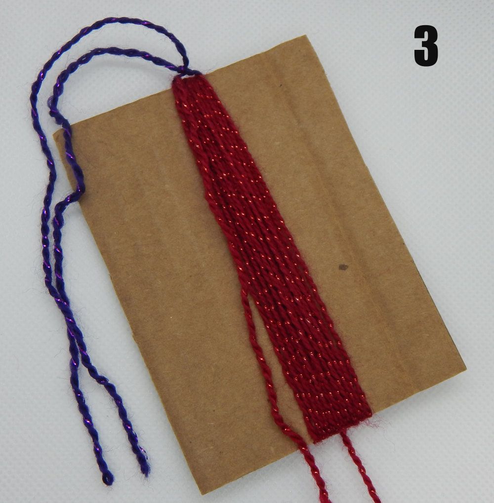 There is a piece of brown cardboard tilted to the left with multiple strands of red yarn wrapped around it lengthwise. There is a purple cord tying the red cords together at the top. There is a 3 in the upper right hand corner of the picture. 