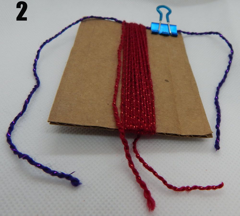 A piece of cardboard has a purple piece of thread attached to it with a blue binder clip on the upper right hand corner. There is multiple strands of red yard wrapped around the rectangle of cardboard. There is a 2 in the upper left hand corner of the picture.
